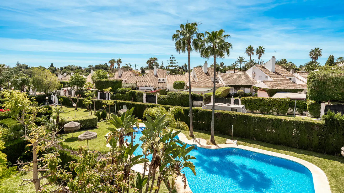 Aerial view of a bright blue swimming pool surrounded by lush green lawns, hedges, and palm trees in a residential area. White houses with terracotta roofs are visible in the background.