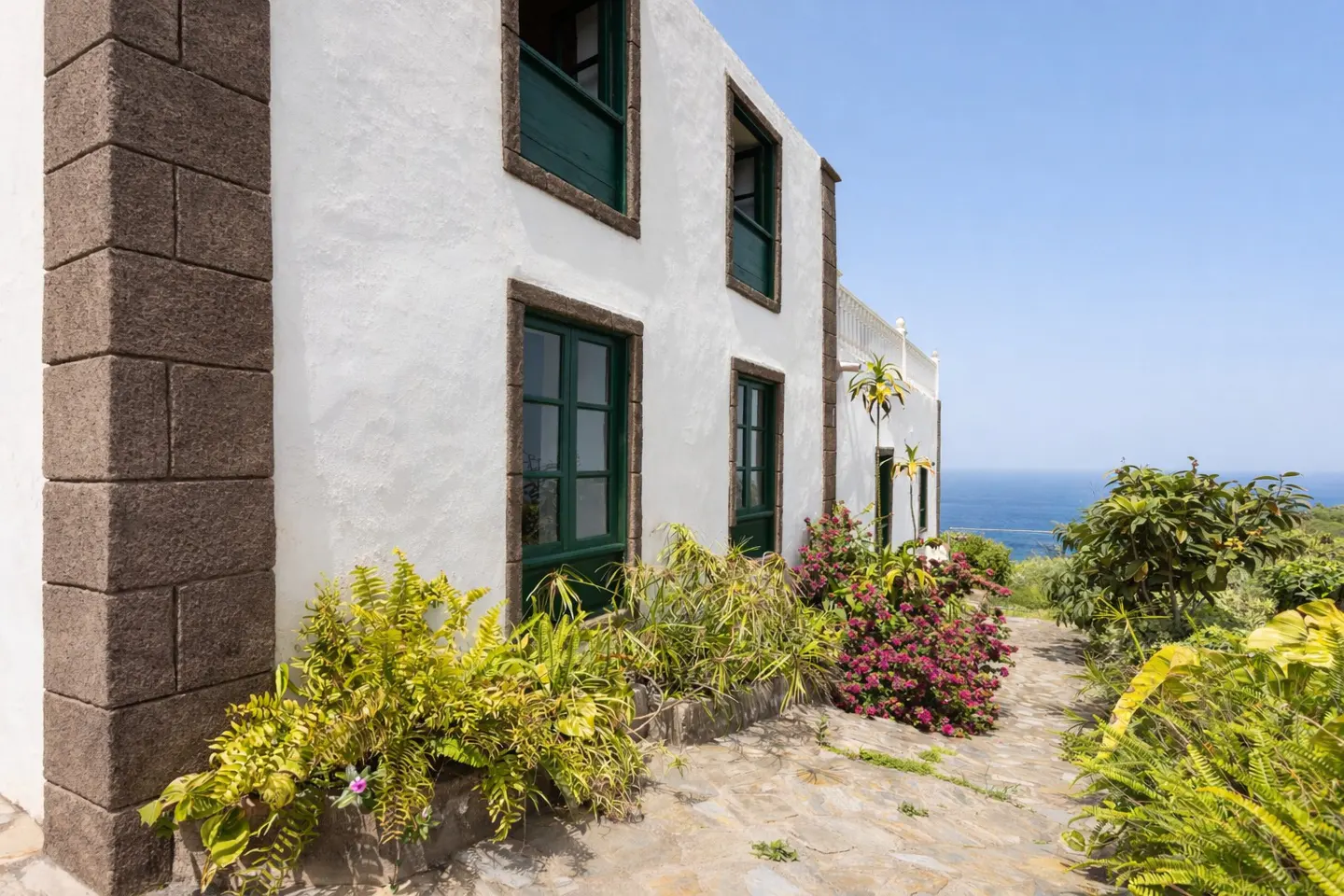 Exterior of a white house with green window frames, stone accents, and lush greenery, overlooking the ocean under a clear blue sky.