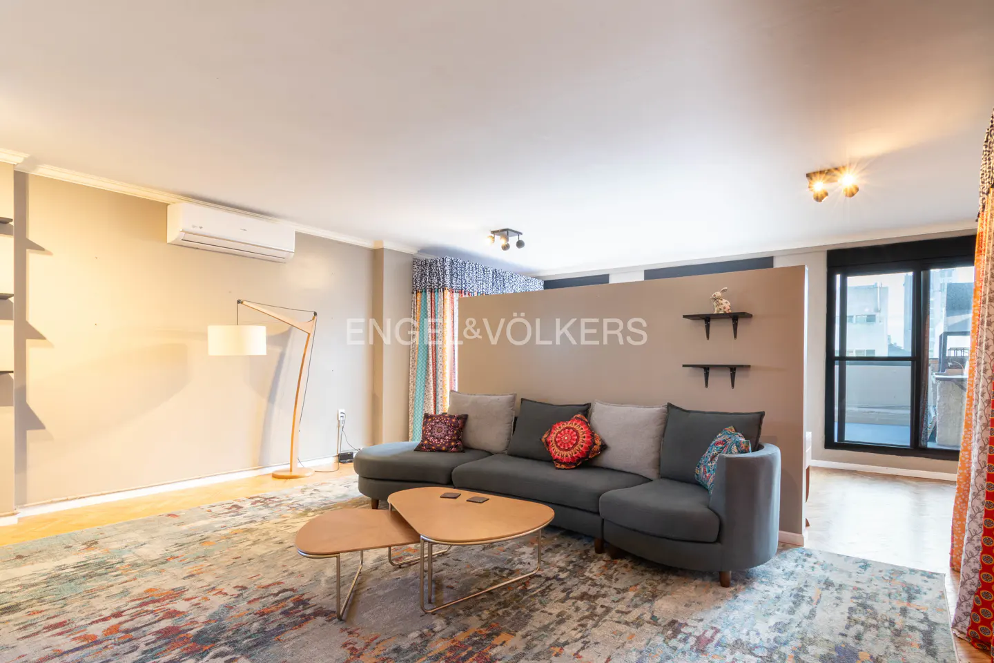 Living room with a gray sectional sofa, two-tiered coffee table, and colorful rug. A floor lamp stands near a wall-mounted air conditioner.