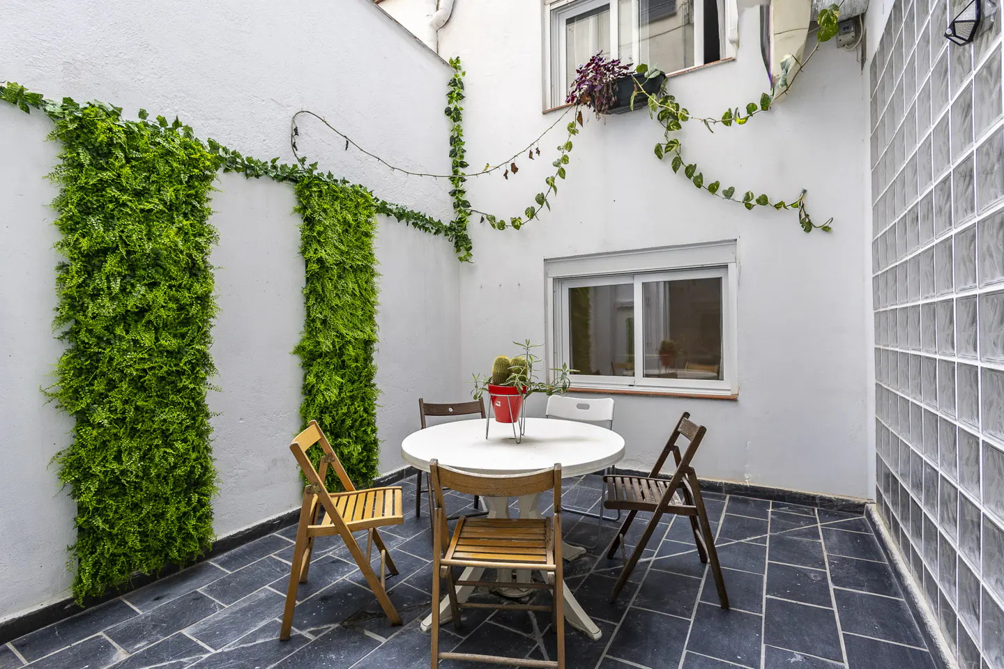 Small patio with a white round table, wooden chairs, vertical green wall panels, and a glass block wall on dark tile floor.