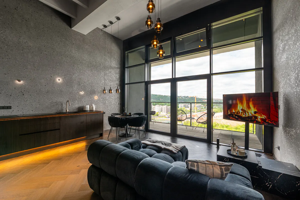 Modern apartment interior with a blue sofa, black kitchen cabinets, and a large window with a city view. A TV displays a fireplace.