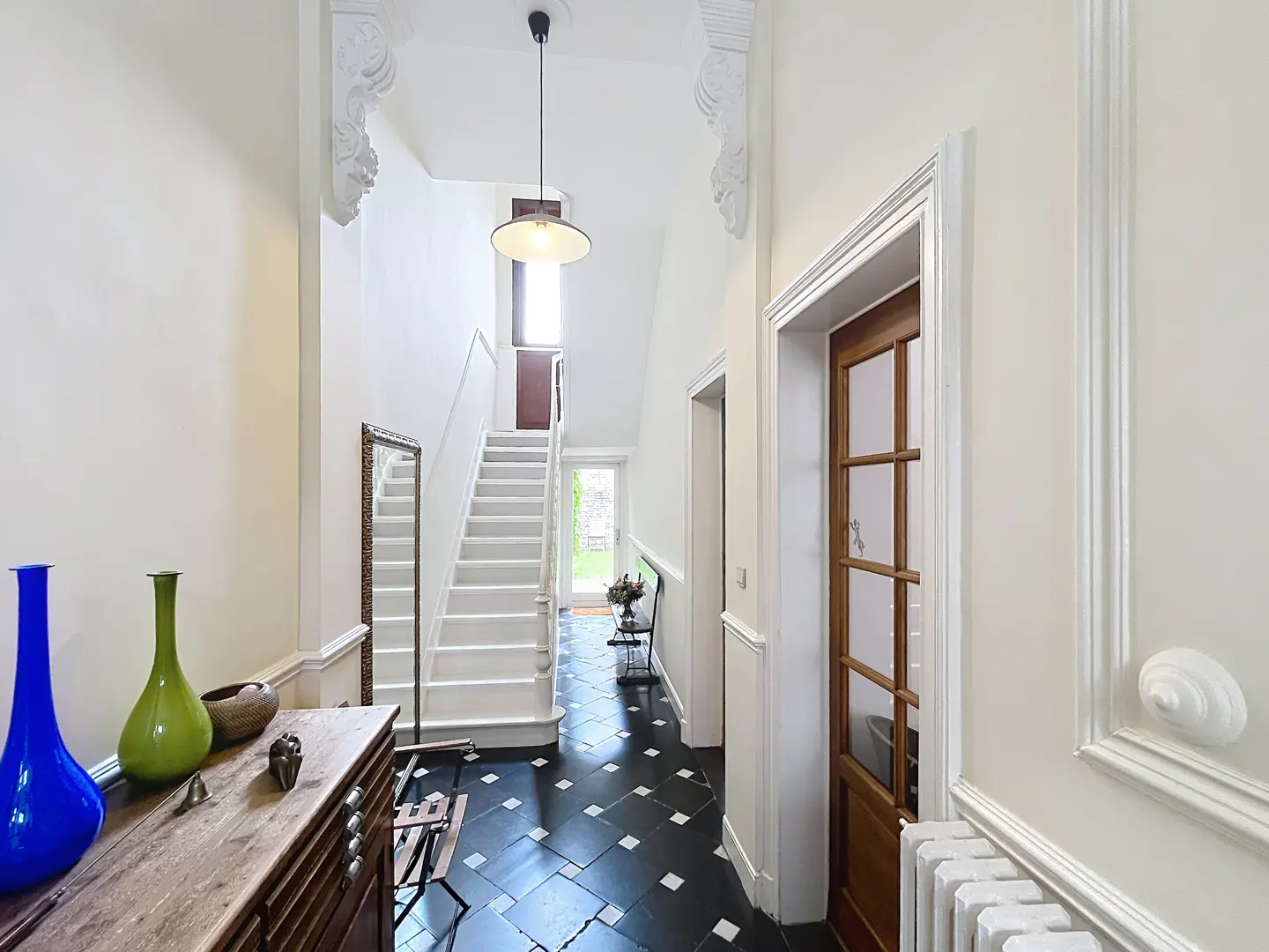Bright hallway with black and white tiled floor, white staircase, wooden console table, and blue and green glass vases.