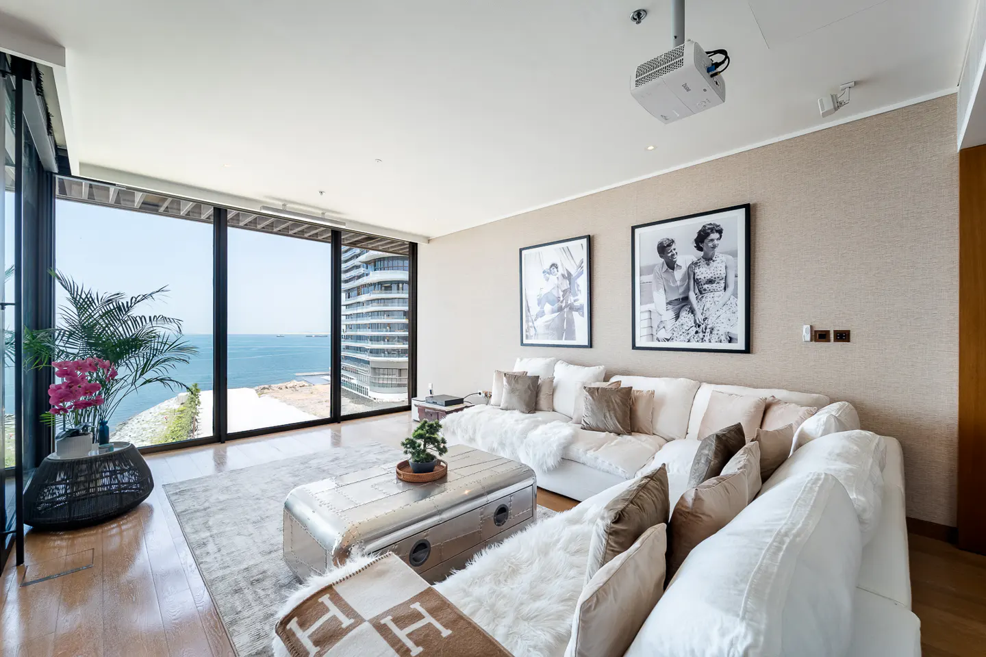 Bright living room with a white sofa, metal coffee table, and large windows overlooking the ocean. Black and white photos hang on the wall.