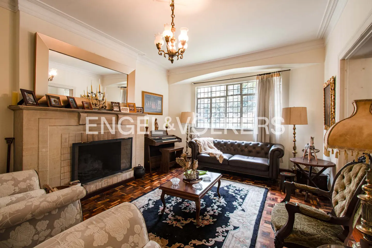 Living room with fireplace, brown leather sofa, piano, and floral rug on a parquet floor. A chandelier hangs from the ceiling.