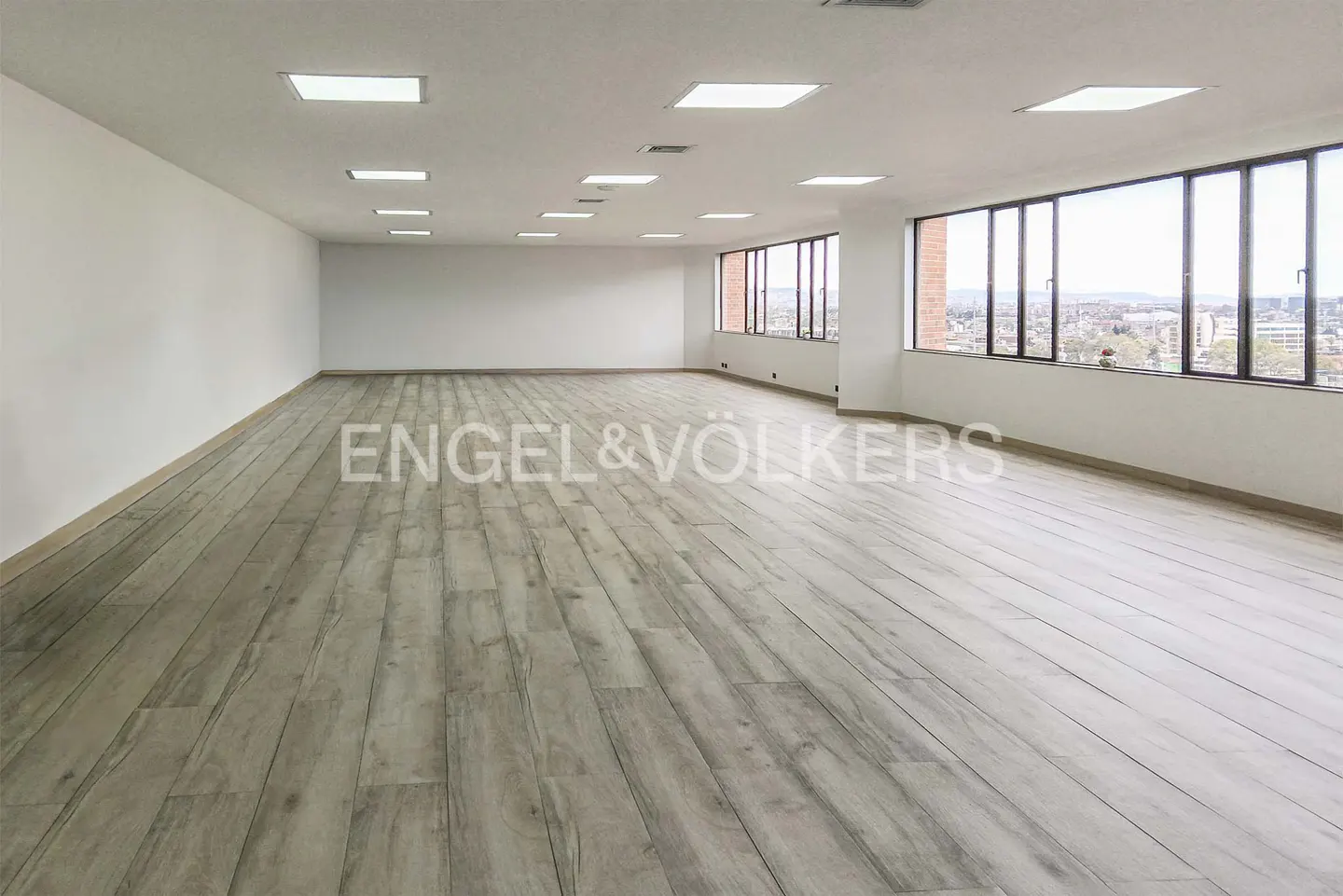 Bright, empty office space with wood floors, white walls, and large windows overlooking a cityscape. Recessed lighting in the ceiling.