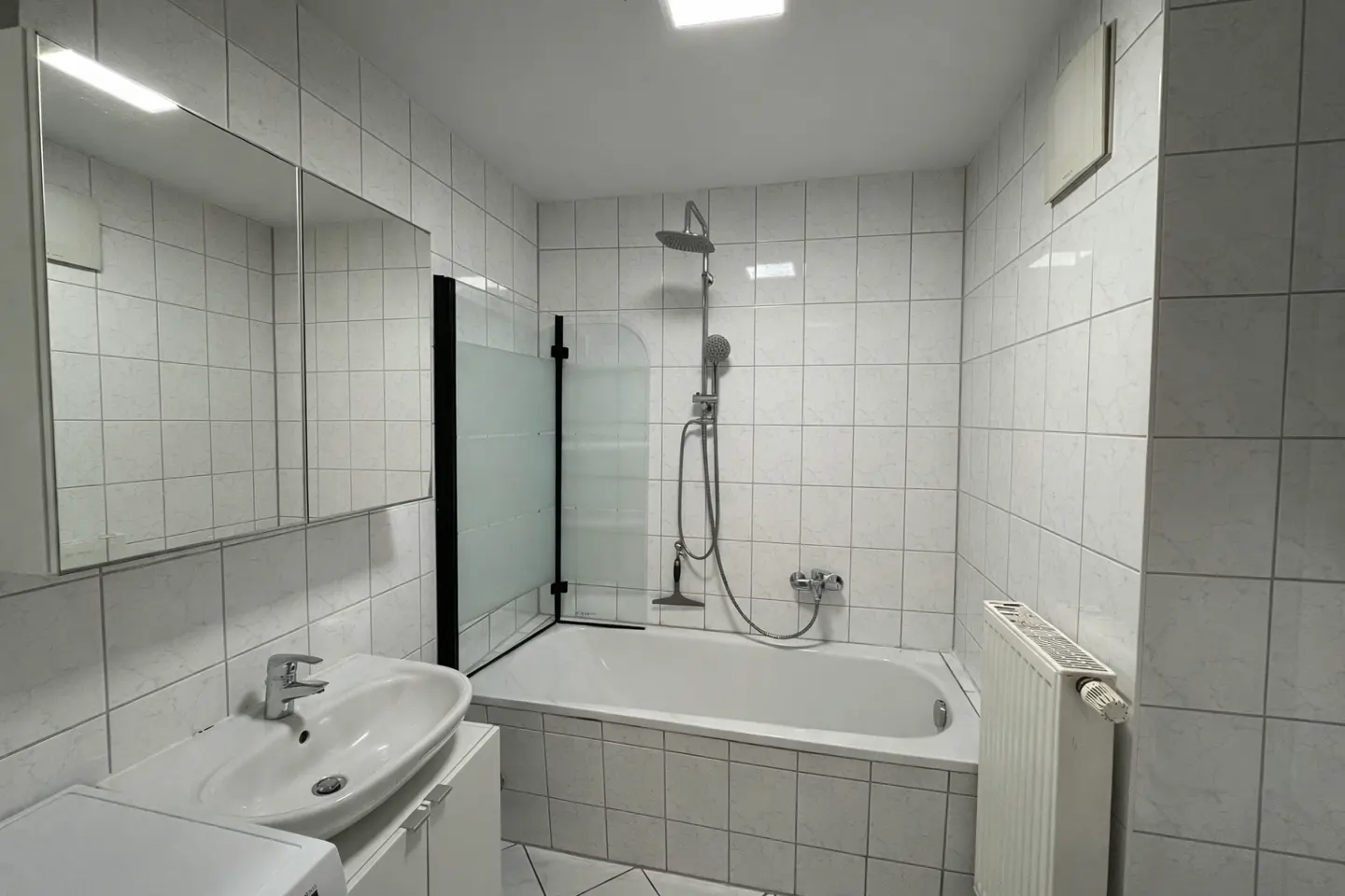 A white tiled bathroom with a sink, mirror, bathtub with a shower head, and a radiator.