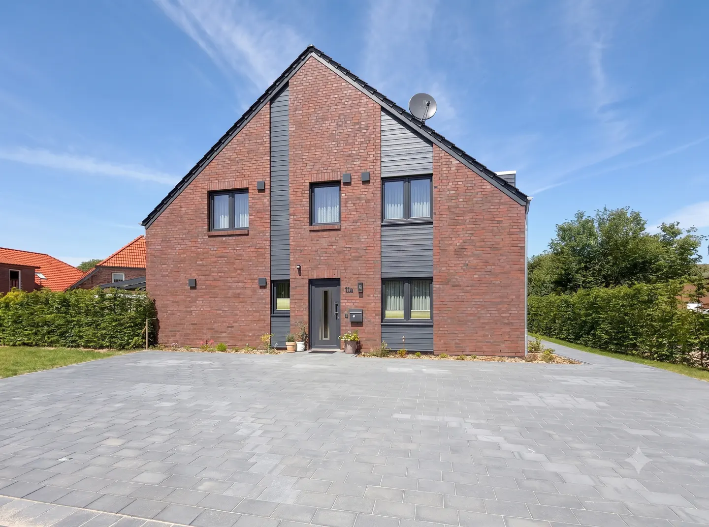Two-story brick house with gray accents, a gray paved driveway, and a satellite dish on the roof against a blue sky.