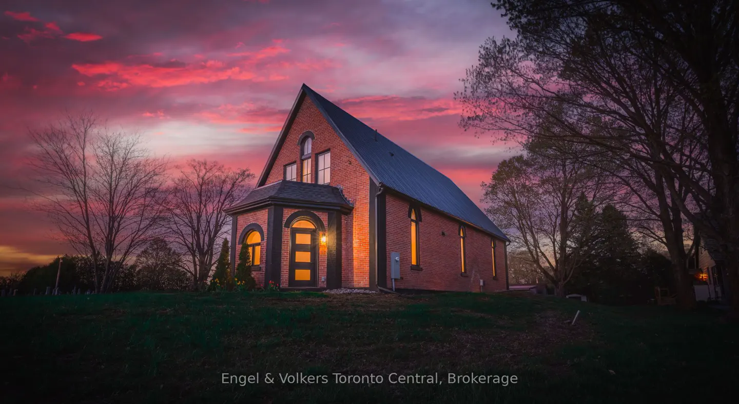 A red brick house with a steep roof glows under a vibrant pink and purple sunset, surrounded by trees on a grassy hill.