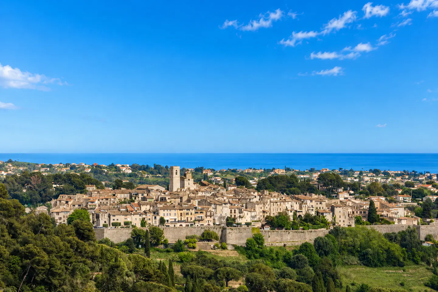 Scenic view of Saint-Paul-de-Vence, France, with stone buildings, a bell tower, and a defensive wall, set against a blue sky and sea.