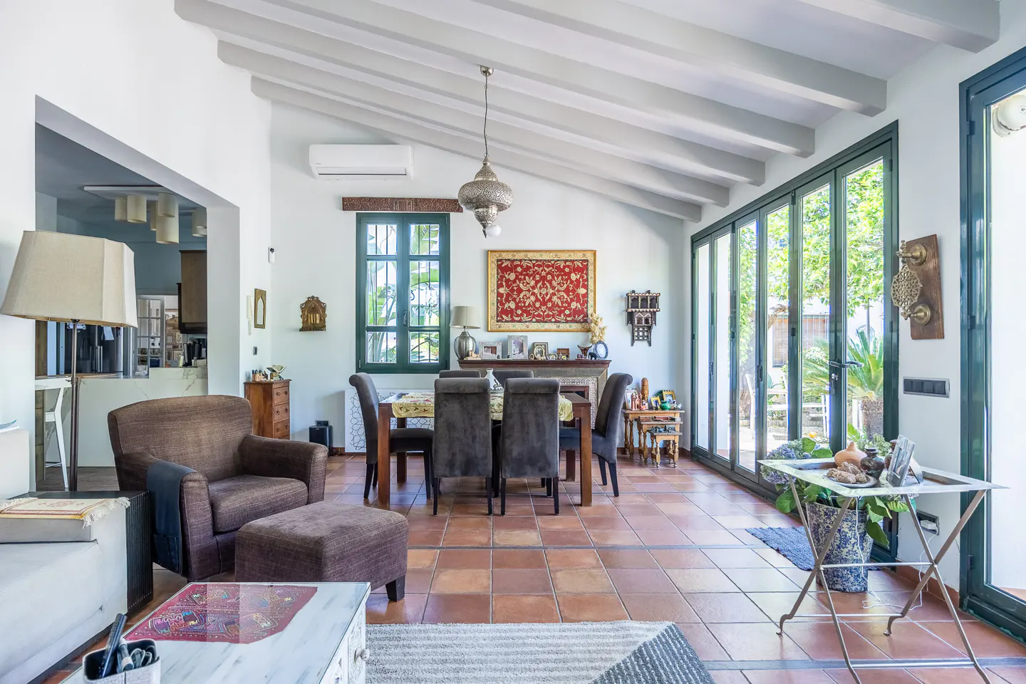 Bright living room with terracotta tile floor, white walls, and exposed beam ceiling. A dining table and brown armchair are visible.