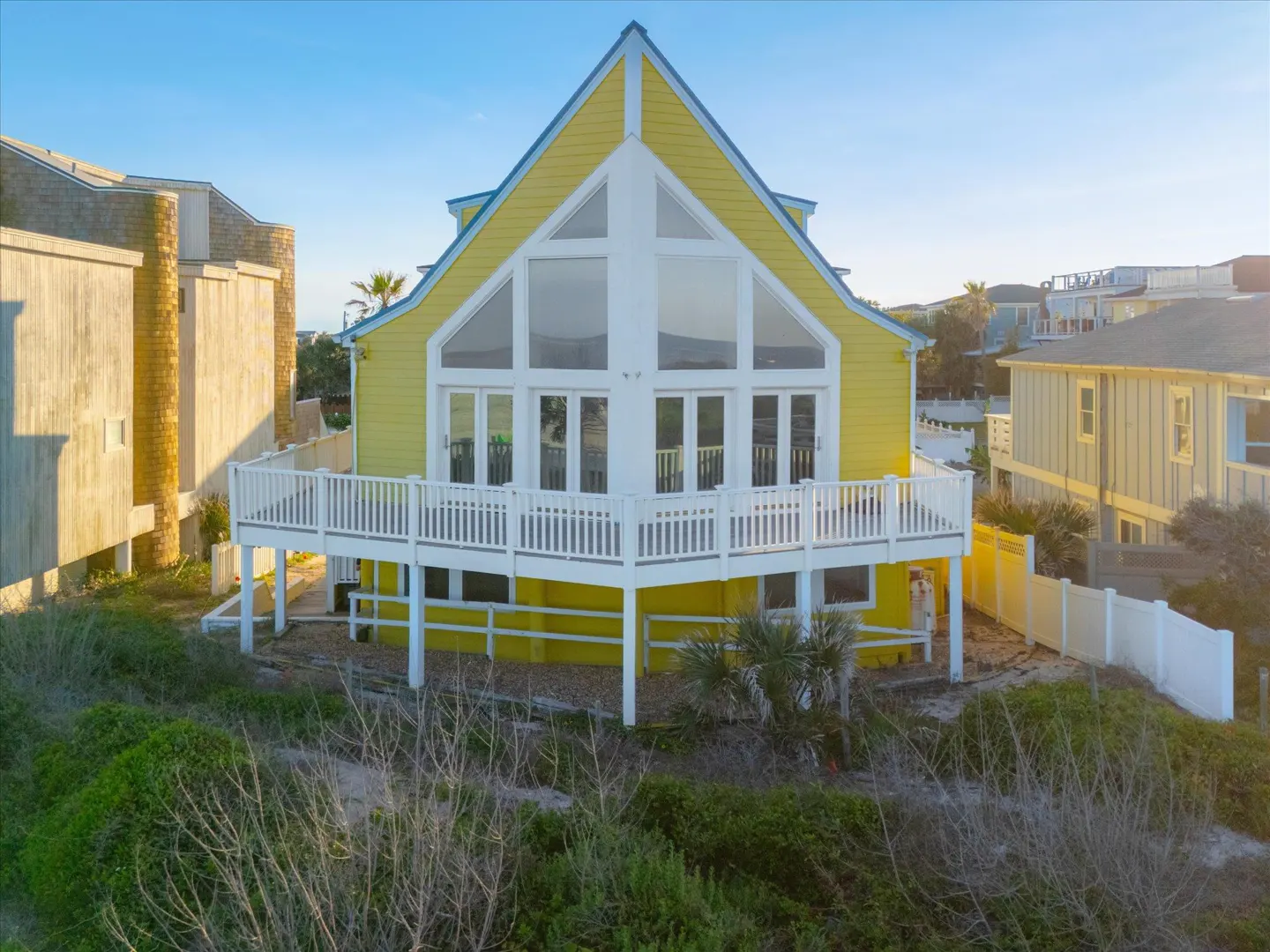 Bright yellow A-frame beach house with a large white deck and floor-to-ceiling windows, surrounded by coastal vegetation.