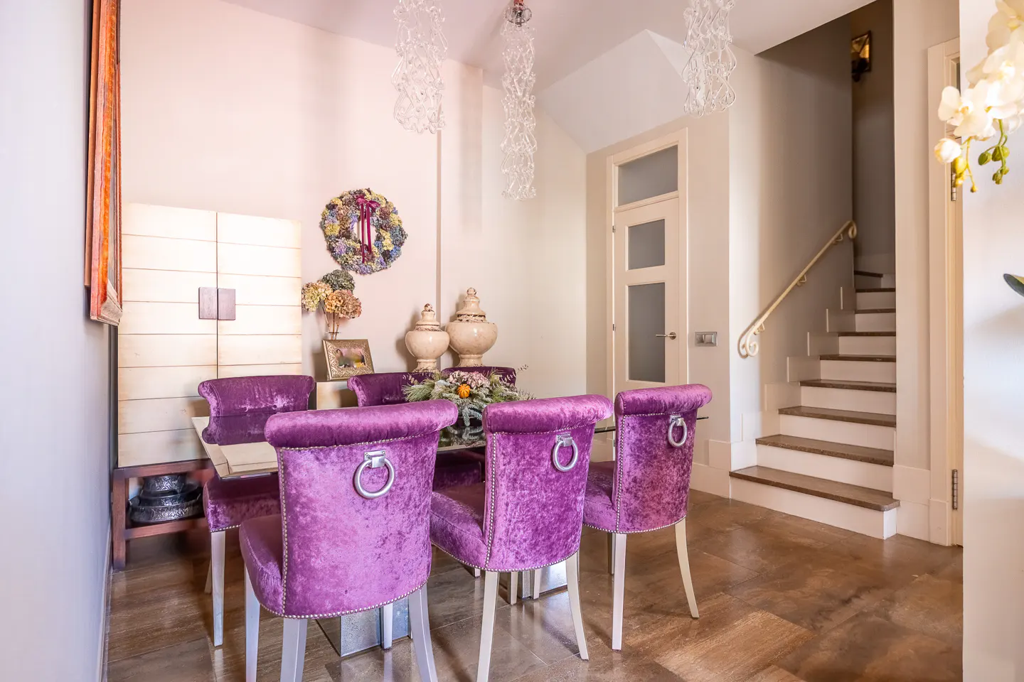 Dining room with purple velvet chairs around a glass table, white cabinet, and staircase in the background.