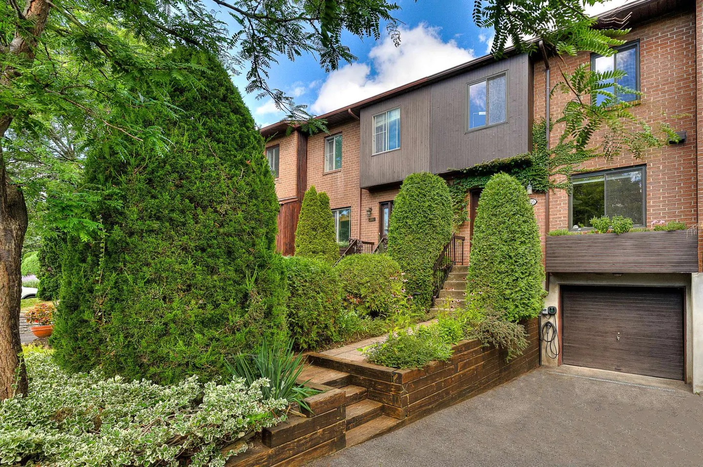 Exterior of a brick townhouse with a brown garage door and lush green landscaping.