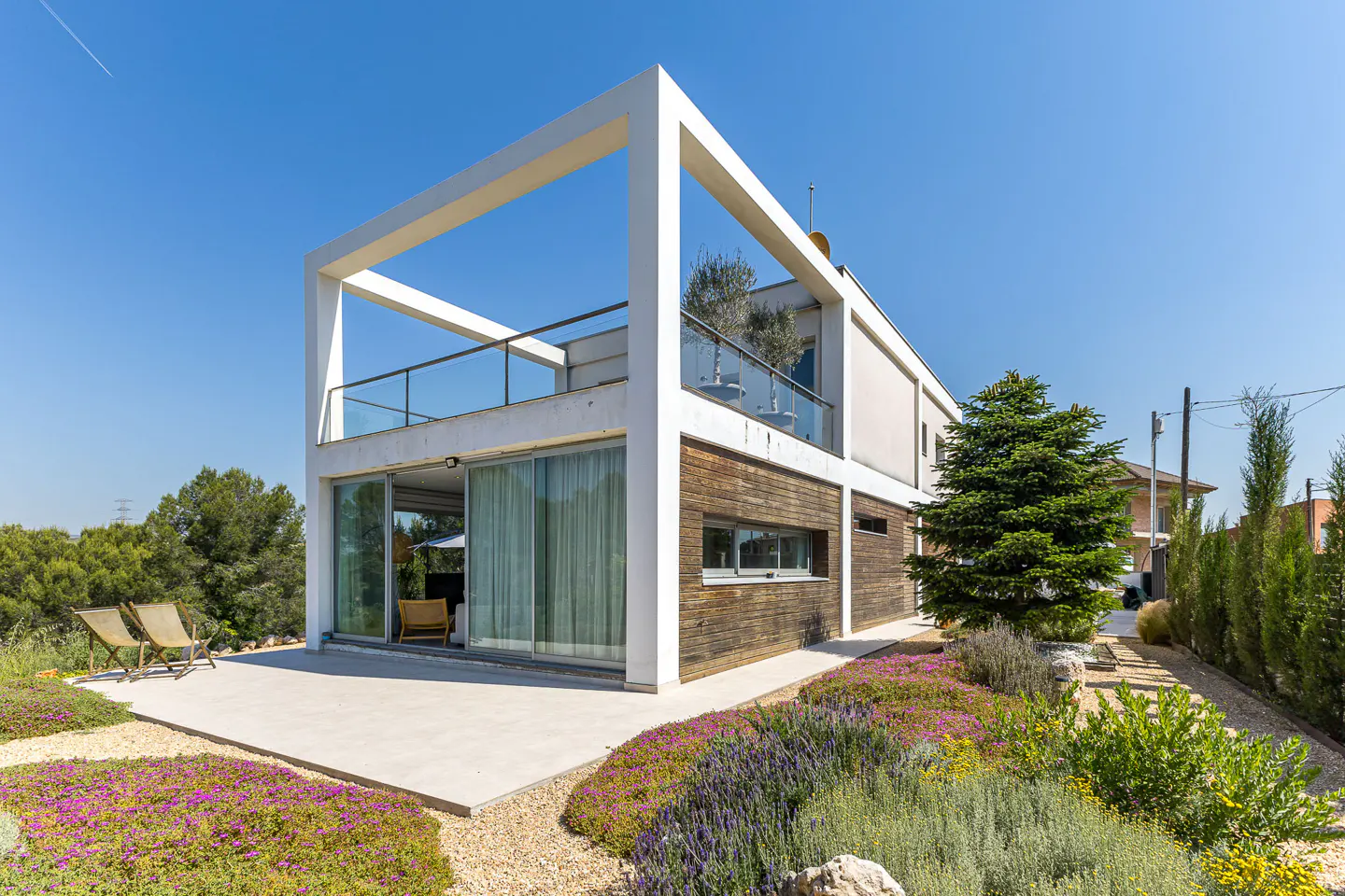 Modern two-story house with white frame, wood siding, and glass balcony. Patio with chairs and colorful flower garden. Blue sky.