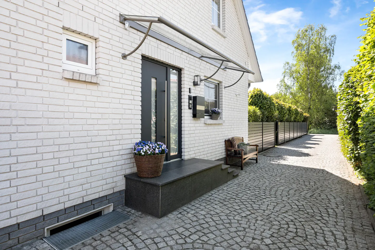 White brick house exterior with a modern dark grey front door, stone steps, a flower basket, and a cobblestone driveway.