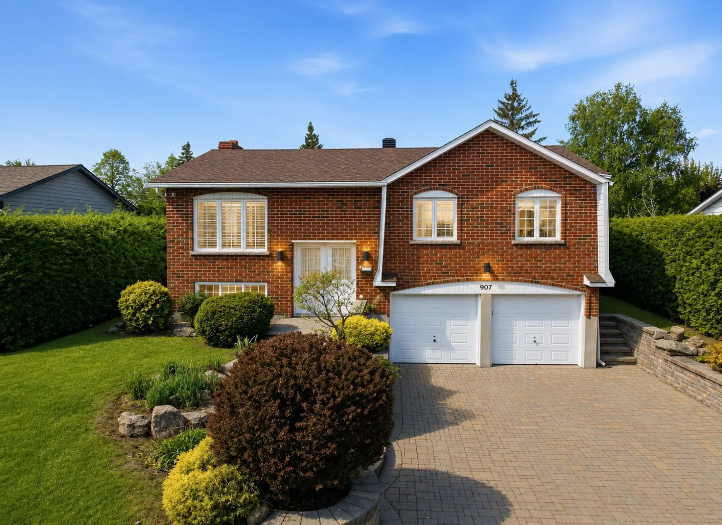 Red brick house with a brown roof, white garage doors, and a paved driveway. Green lawn and bushes surround the house.