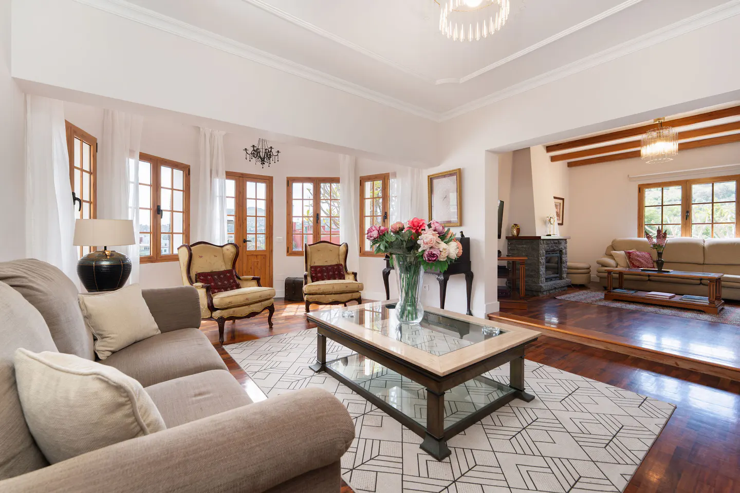 Bright living room with a beige sofa, two armchairs, a glass coffee table with flowers, and wood-framed windows.