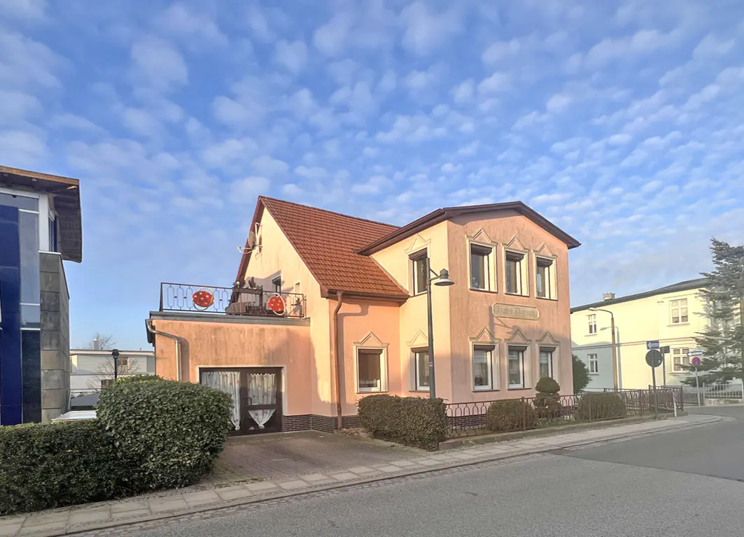 A two-story pink house with a red roof and a balcony with red decorations. The sky is blue with white clouds.