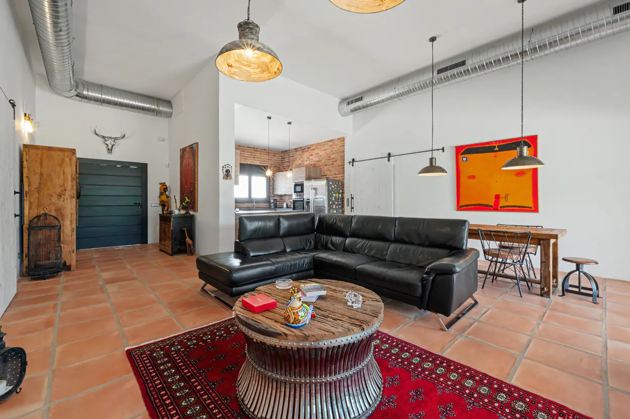 Industrial-style living room with black leather sectional, rustic round coffee table, red rug, and open kitchen beyond.