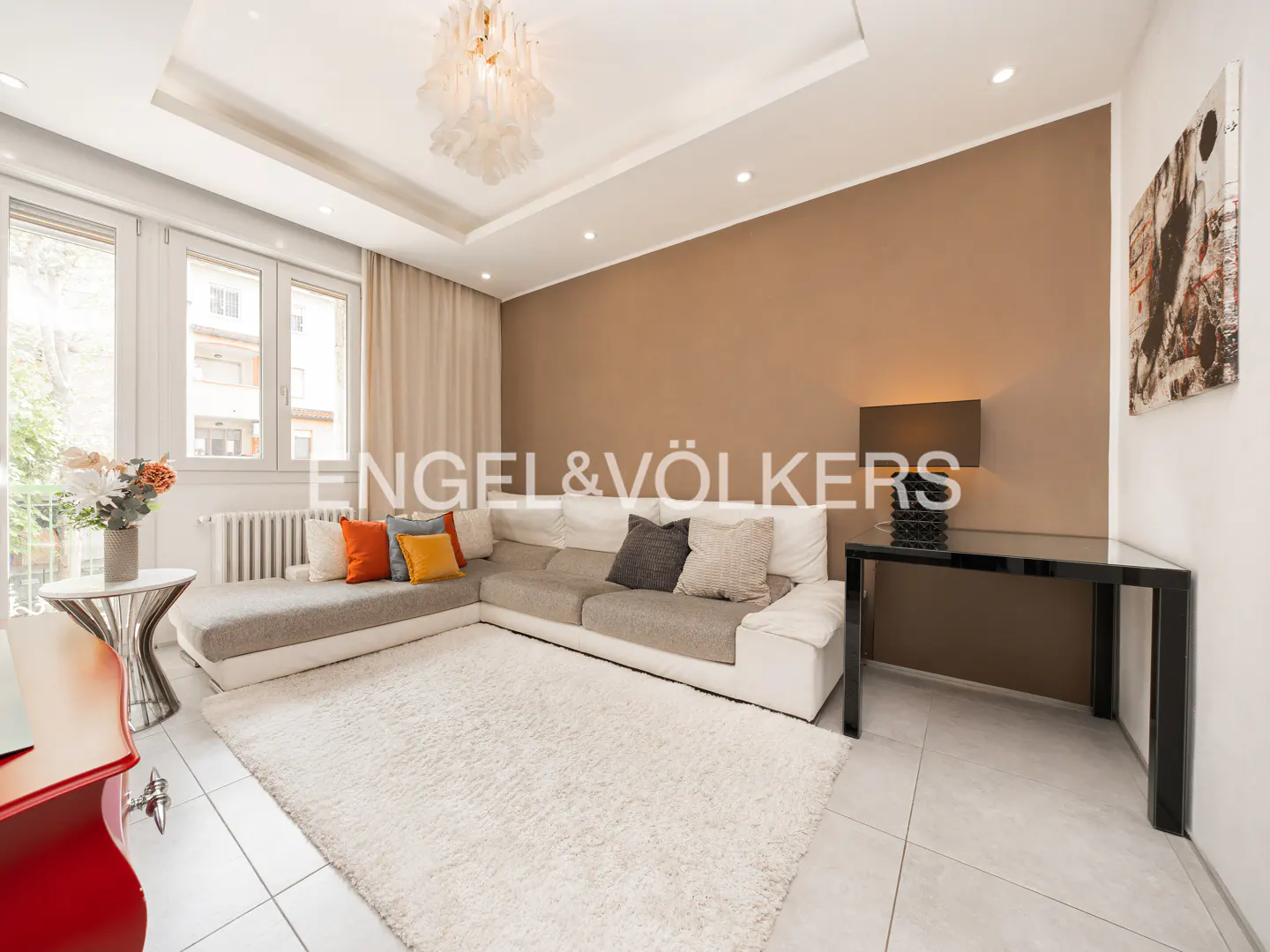 Living room with white L-shaped sofa, fluffy rug, black table, and tan accent wall. Engel & Völkers logo visible.