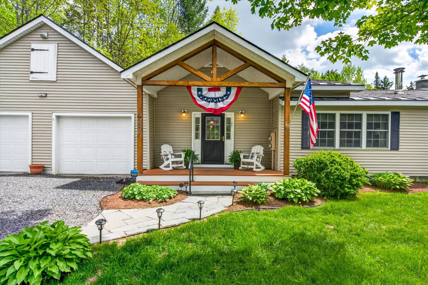 Tan house with a covered porch, two white rocking chairs, American flag, and a stone walkway leading to the front door.