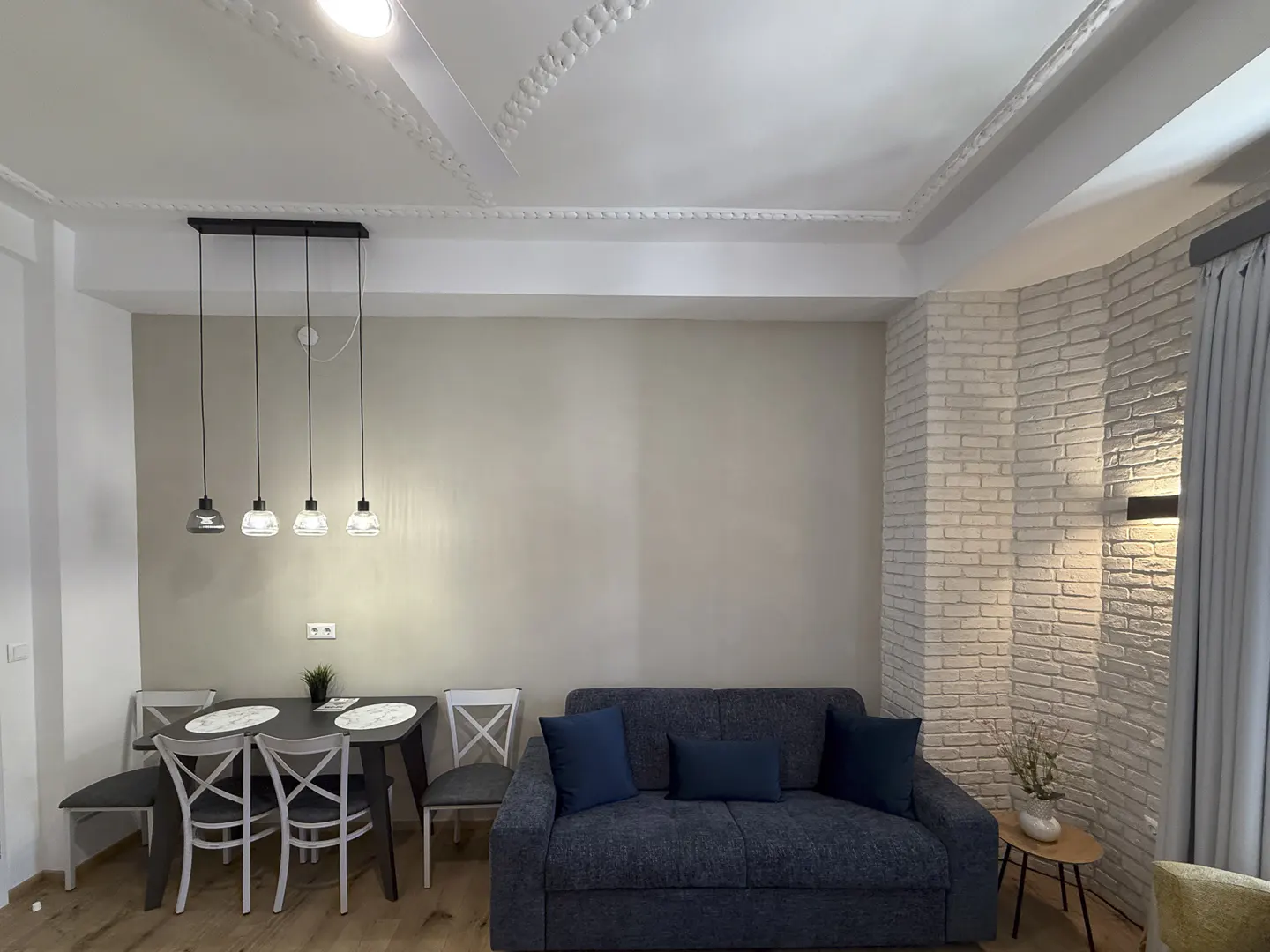 Living room with a blue sofa, dining table with four chairs, and a brick wall accent. Four pendant lights hang above the table.