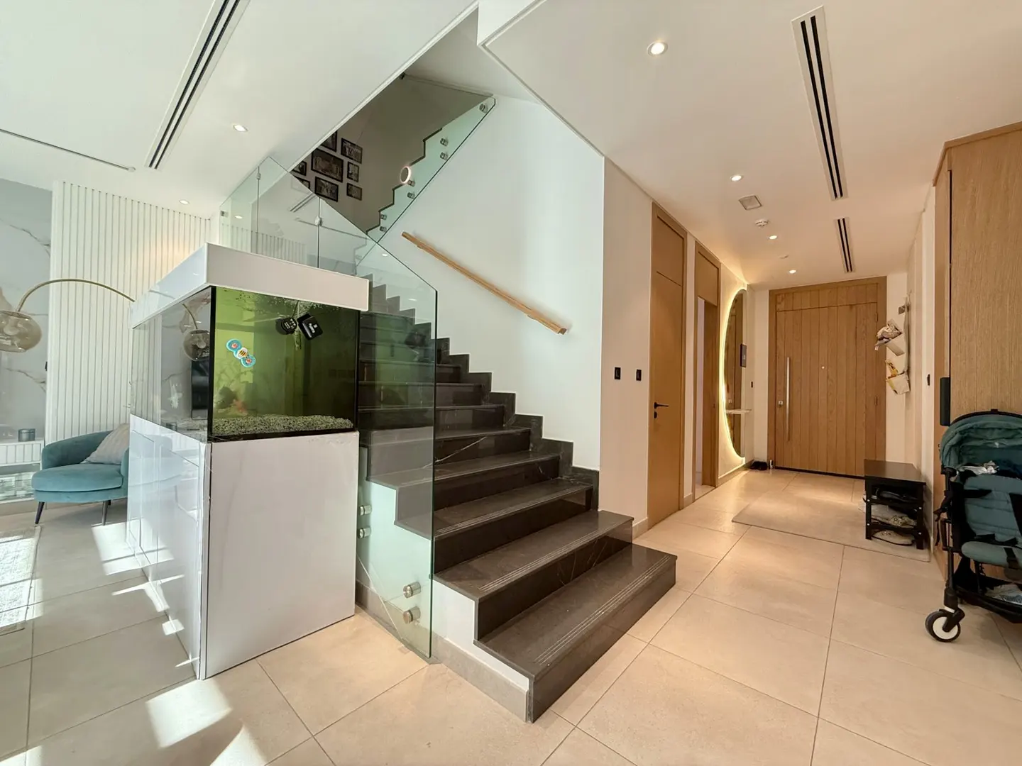 A modern home foyer with a staircase, aquarium, and light wood accents. The stairs have glass railings. A round mirror hangs near the front door.
