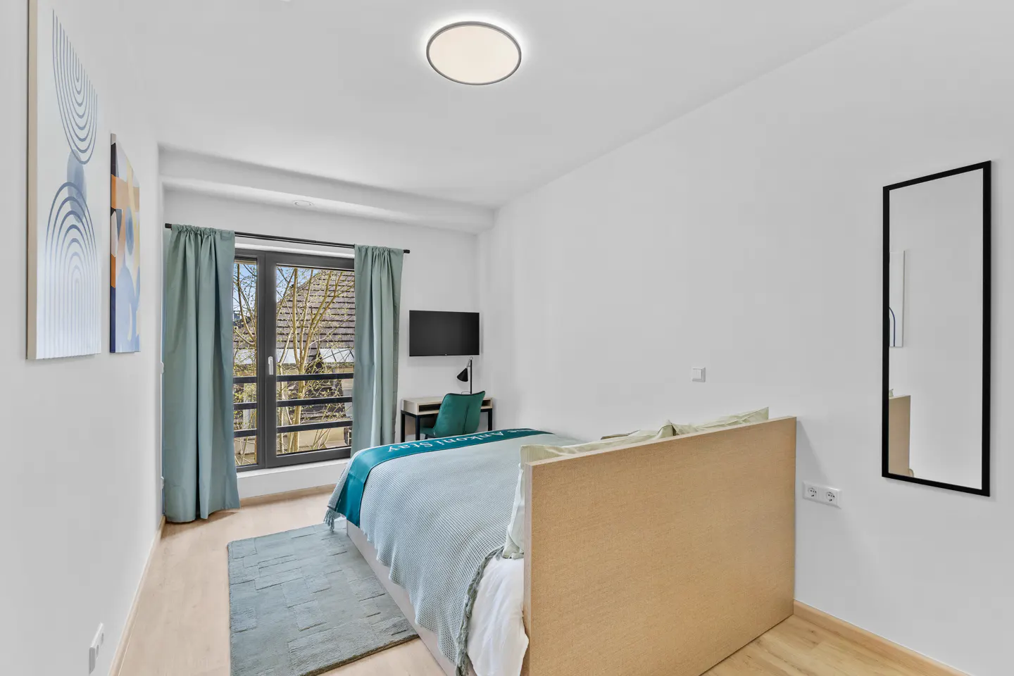 Bedroom with a bed, blue rug, curtains, and a black framed mirror on a white wall. A desk and TV are near the window.