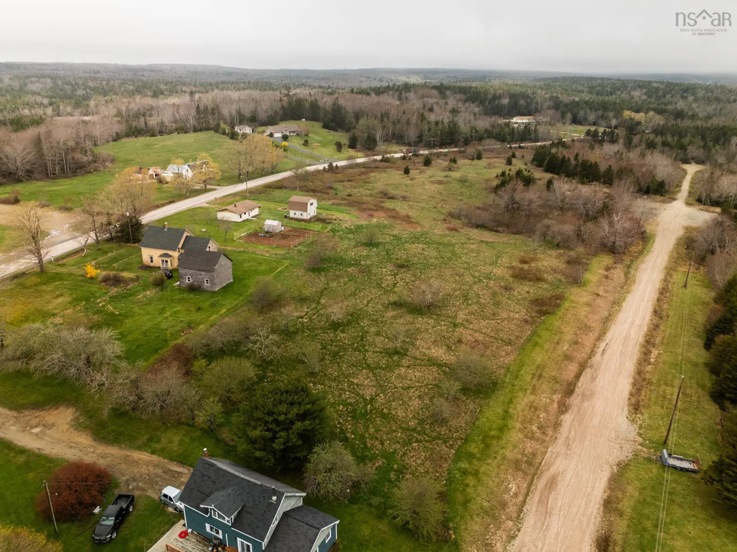 Aerial view of a rural landscape with houses, fields, and a dirt road leading into a forest.