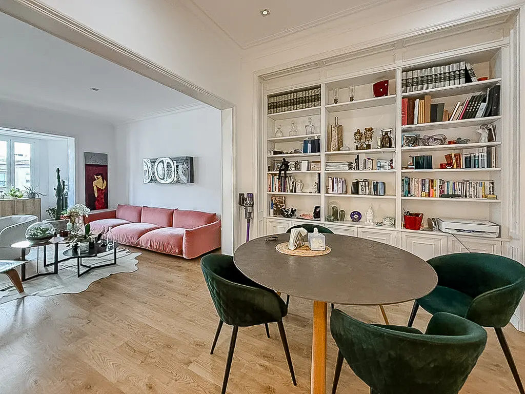 Dining area with a round table and dark green chairs, opening into a living room with a pink sofa and built-in bookshelves.