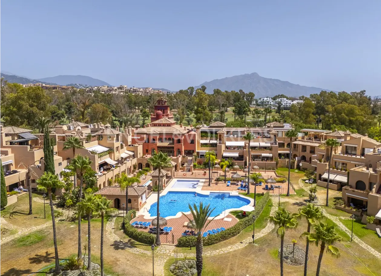 Aerial view of a resort with a blue pool, tan buildings, palm trees, and mountains in the background.