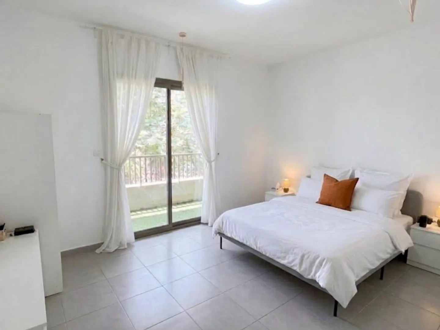 A bright bedroom with white walls, a bed with white linens and an orange pillow, and a sliding glass door to a balcony.