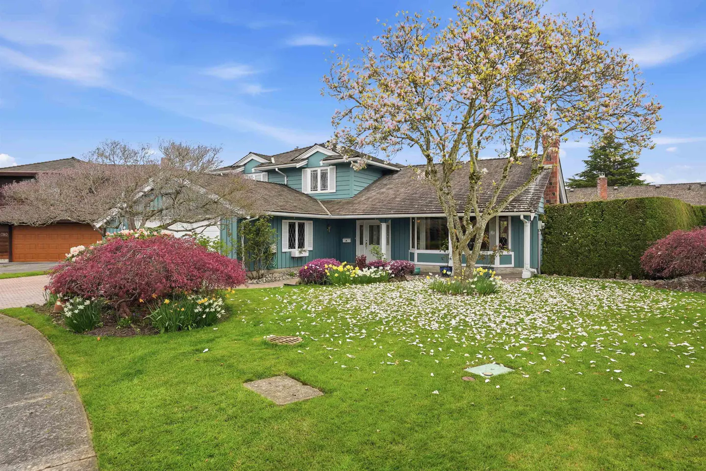 Exterior of a teal house with a brown roof, surrounded by colorful flowers and a tree with white blossoms.