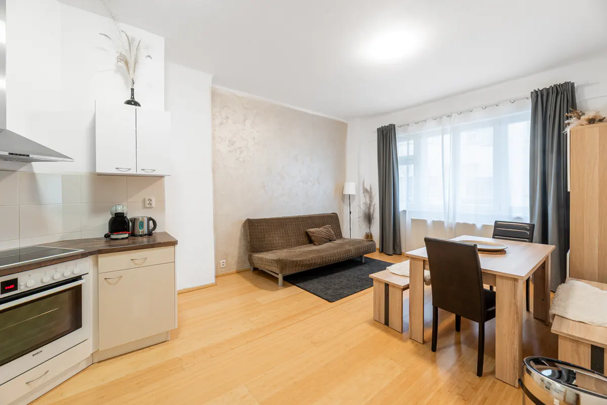Bright apartment interior with wood floors, a brown futon, a dining table with chairs, and a kitchen area with white cabinets.
