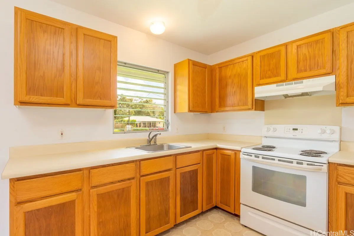 A kitchen with light wood cabinets, white countertops, and a white electric stove. A window with blinds looks out onto a green yard.