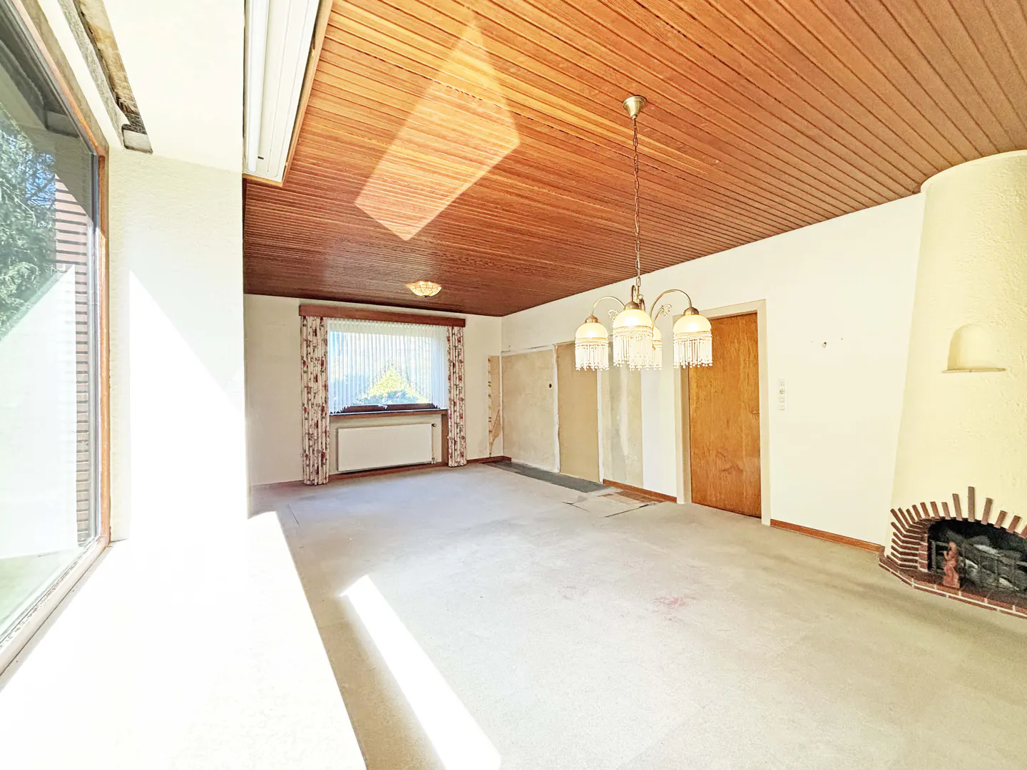 A bright, empty room with a wood-paneled ceiling, a chandelier, and a fireplace. A large window lets in sunlight.