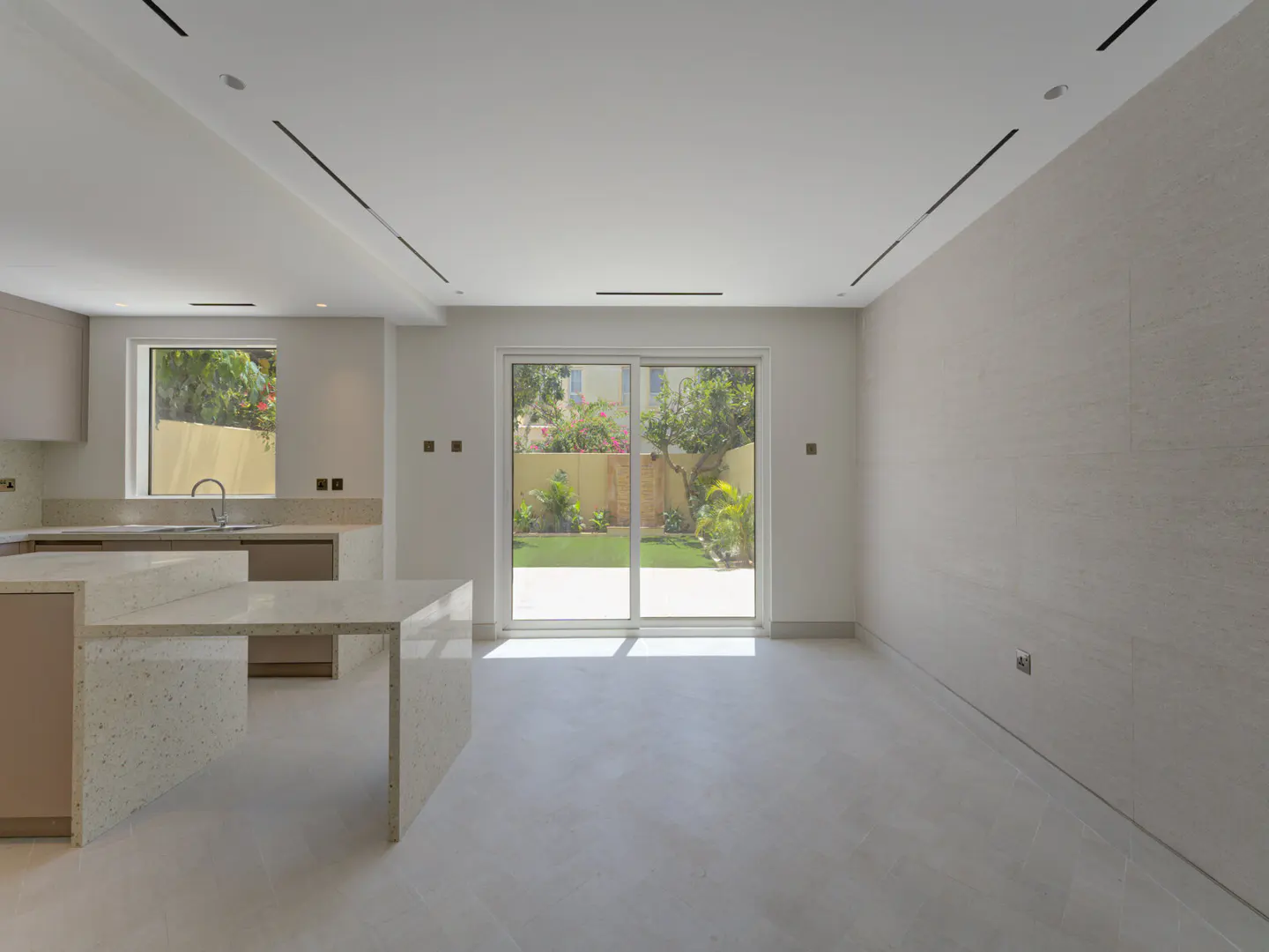 A bright, modern kitchen and living area with white walls, light gray floors, and a sliding glass door to a green backyard.