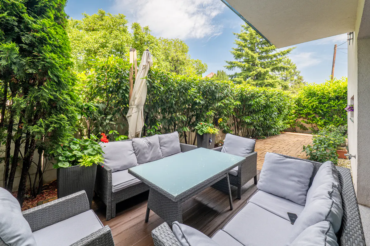 Outdoor patio with gray wicker furniture, including a table and sectional sofa, surrounded by lush green plants and trees.