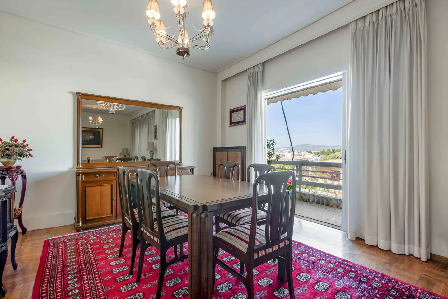 A dining room with a wooden table, chairs, a red rug, and a balcony with a view.