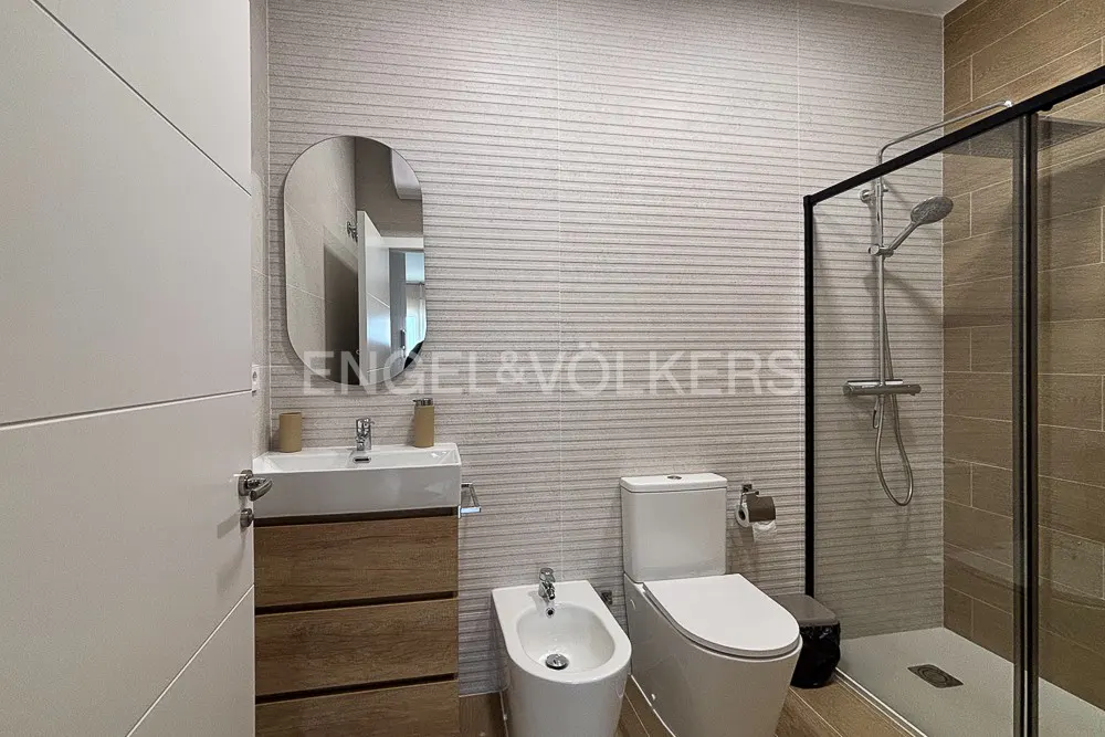Bathroom with white toilet, bidet, sink with wood cabinet, and glass shower. Gray horizontal tile on wall.