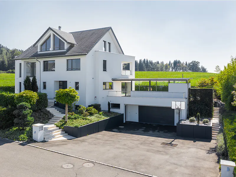 Modern white house with a gray roof, a dark gray garage door, and a basketball hoop in the driveway. Green grass and trees in the background.