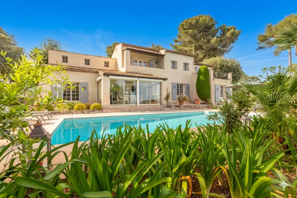 A tan two-story house with a red tile roof overlooks a blue swimming pool surrounded by green plants.