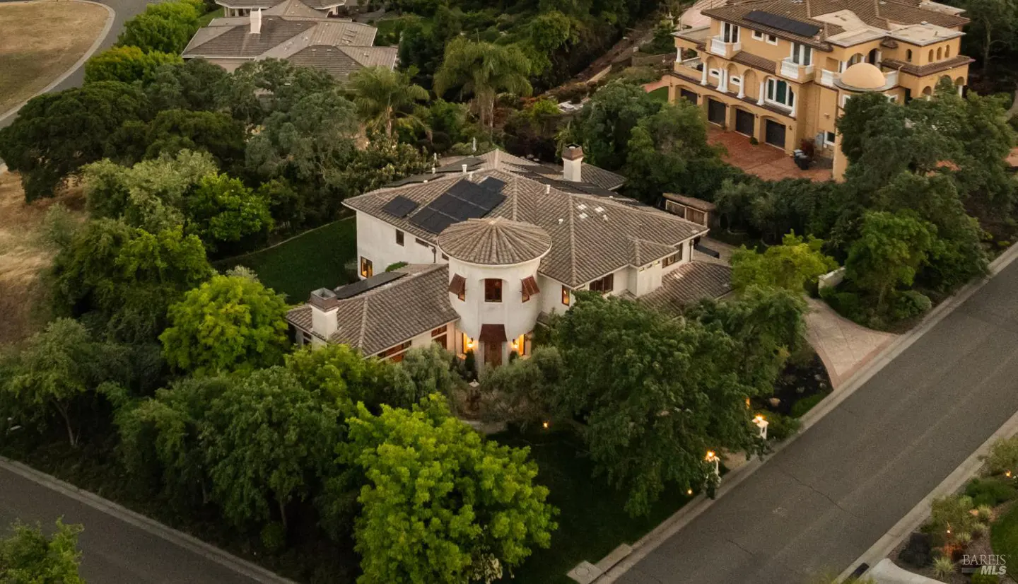 Aerial view of a large, two-story white house with a turret, surrounded by green trees and other houses. Solar panels are visible on the roof.