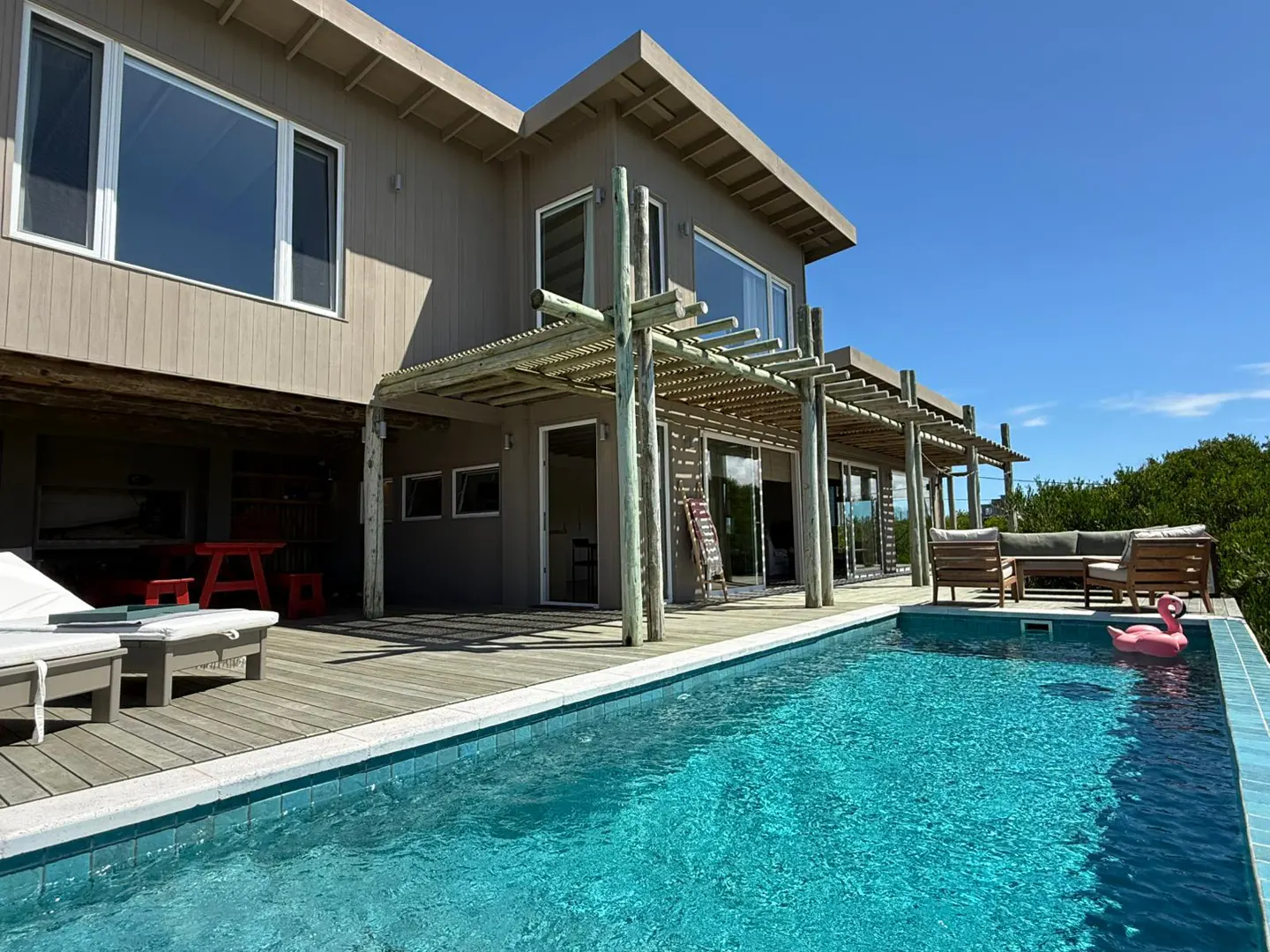 Modern home exterior with a pool. The house is beige with large windows and a wooden pergola. A pink flamingo float is in the pool.