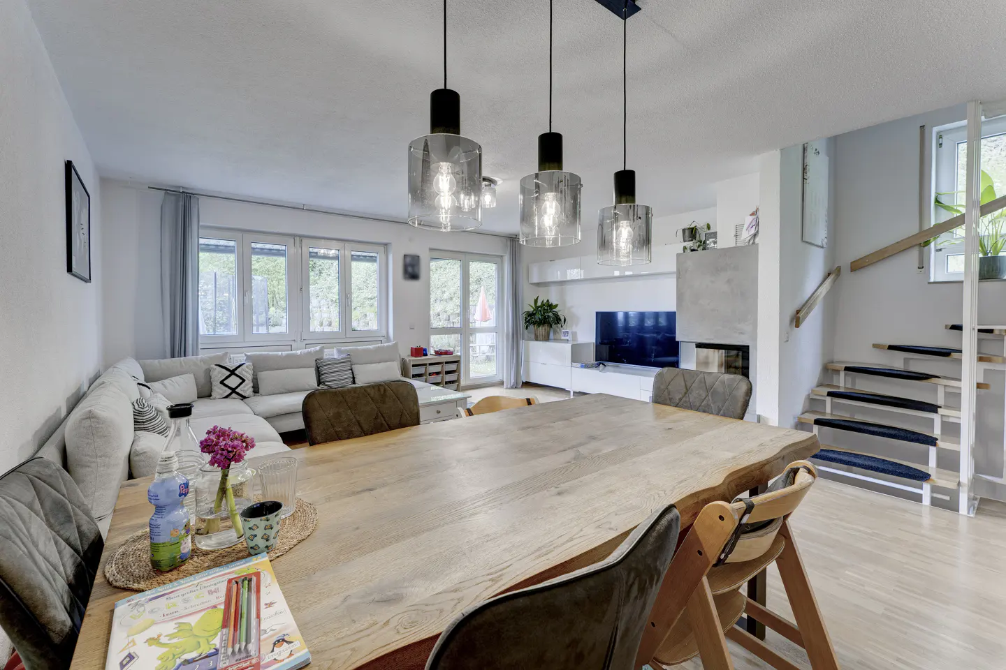 Bright, open-concept living and dining area with a large wooden table, modern lighting, and a comfortable white sofa. Stairs lead to the upper level.