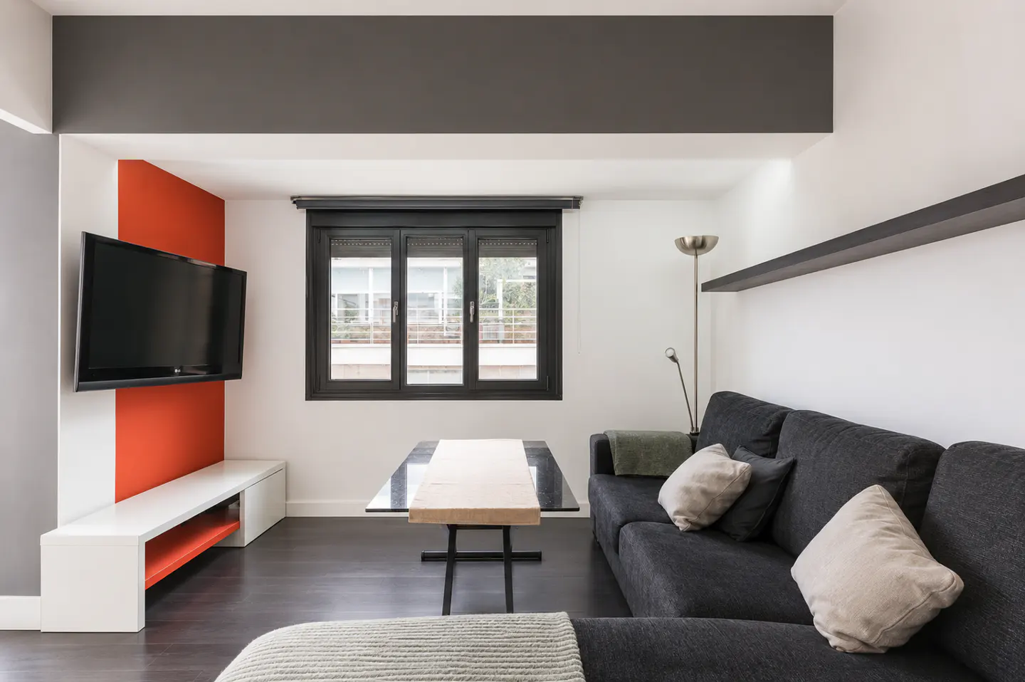 Living room with a gray sofa, coffee table, and TV on an orange accent wall. A window with black frames is centered.