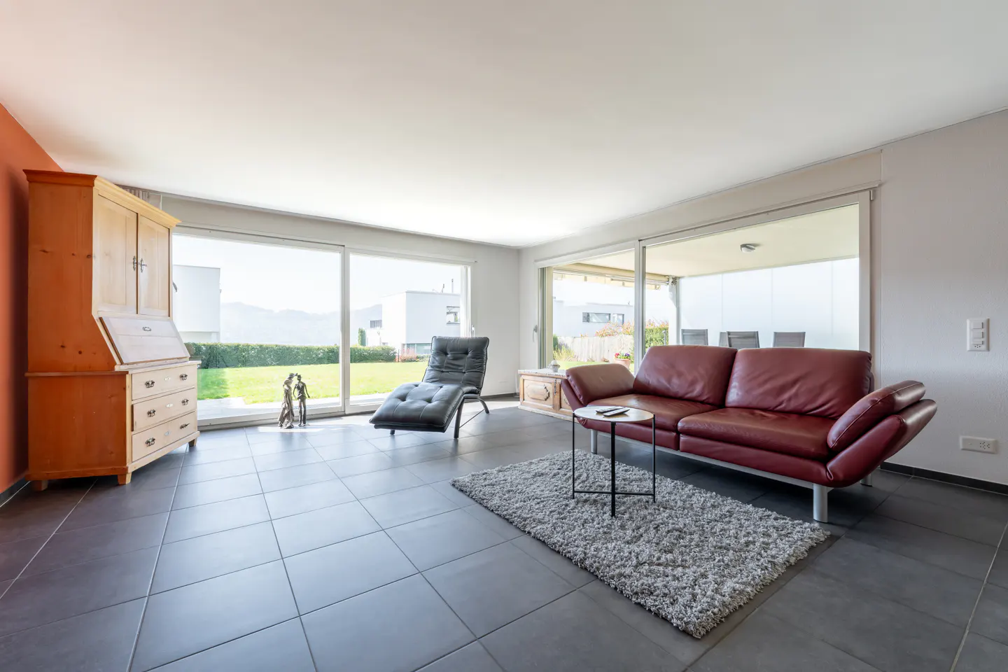 Bright living room with gray tile floor, red leather sofa, chaise lounge, and wooden cabinet. Large windows overlook a green lawn.