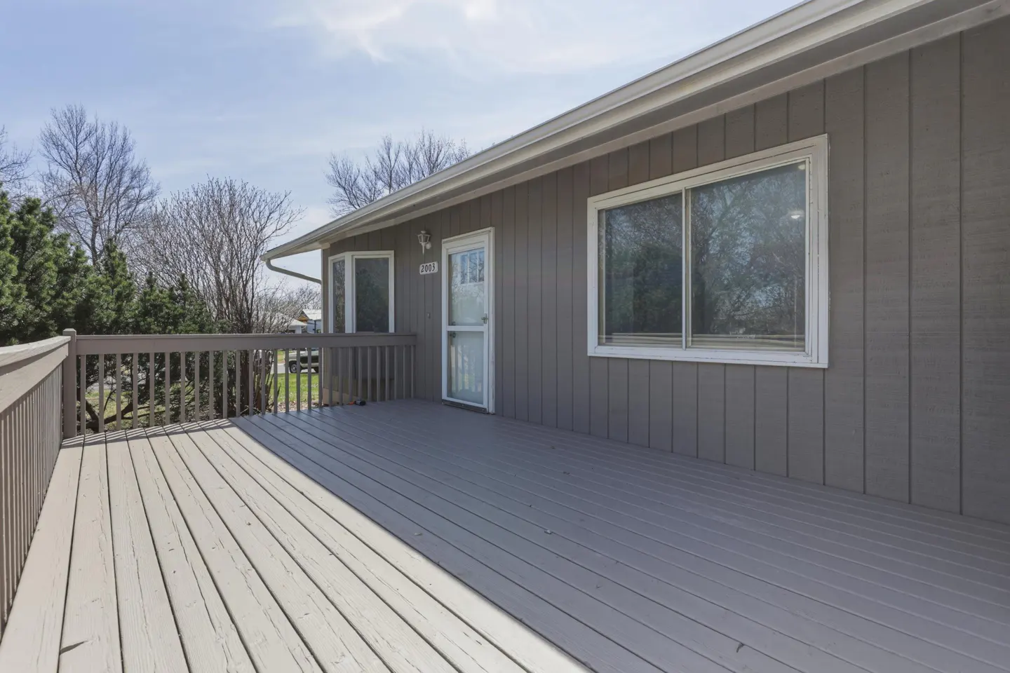 Exterior view of a gray house with a large wooden deck, white door, and windows under a blue sky.