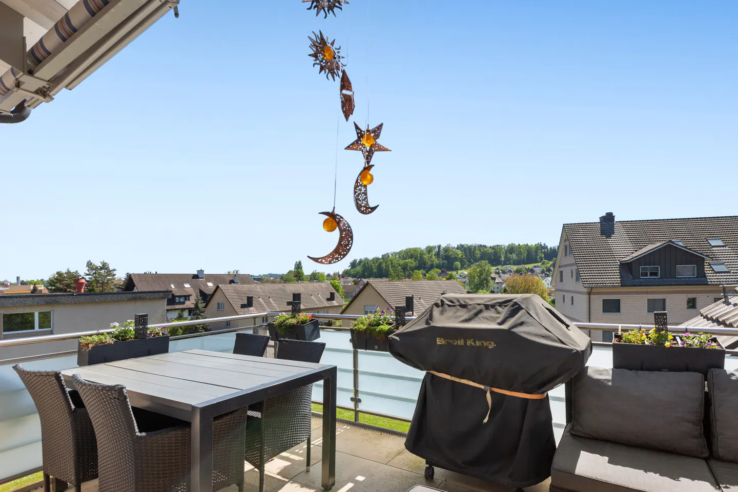 Balcony view with table, chairs, covered grill, and sofa. Decorative sun, moon, and star mobile hangs above. Rooftops and blue sky in background.