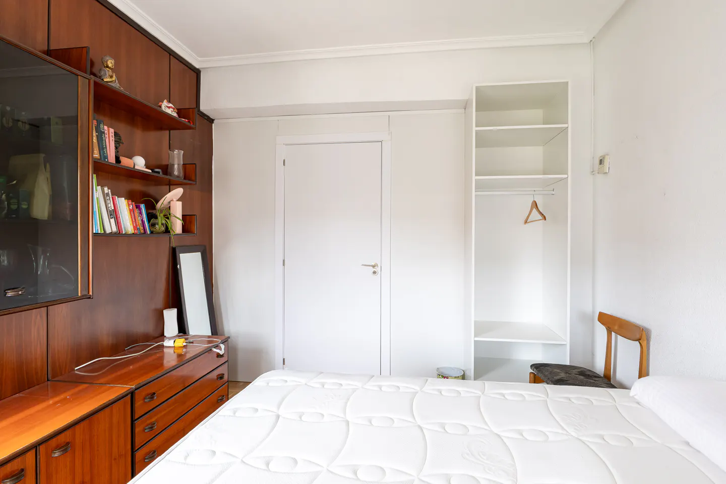 Bedroom with a white quilted bed, brown wood dresser, shelves with books, and a white closet with a hanger.